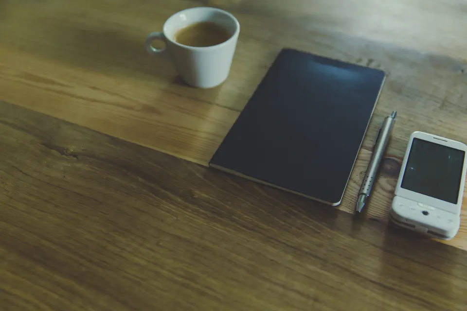 A phone, a closed notebook and coffee on a wooden table.