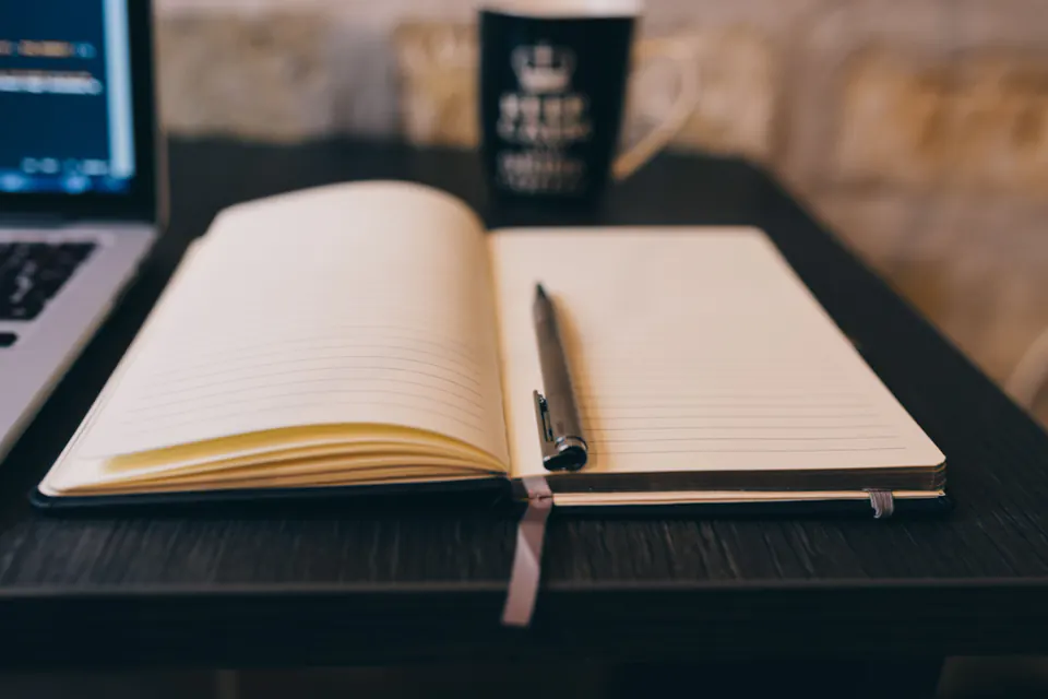 An open notebook with a pen on a wooden desk beside a laptop.